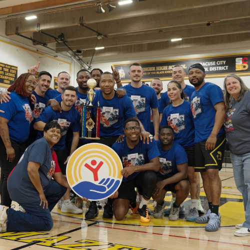 Group of smiling basketball players and coaches posing with a trophy on a gym floor.